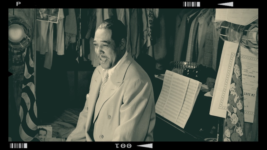 Duke Ellington sits in a back-stage artist's room with a white suit on
