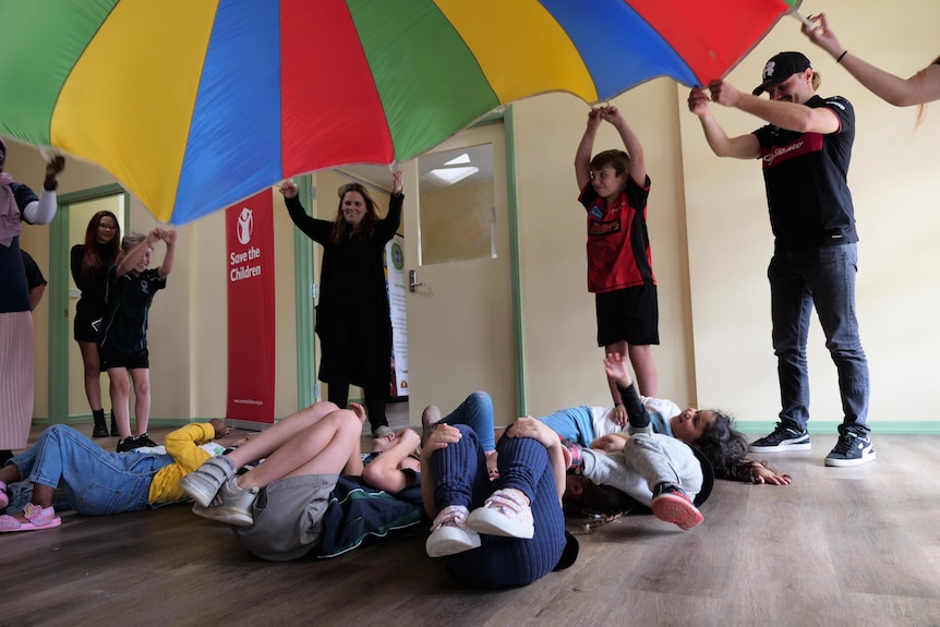A group of adults hold a parachute above a group of kids lying on a floor.