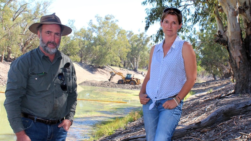 A man and a woman by a river with earthmoving equipment seemingly making a dam in the background.