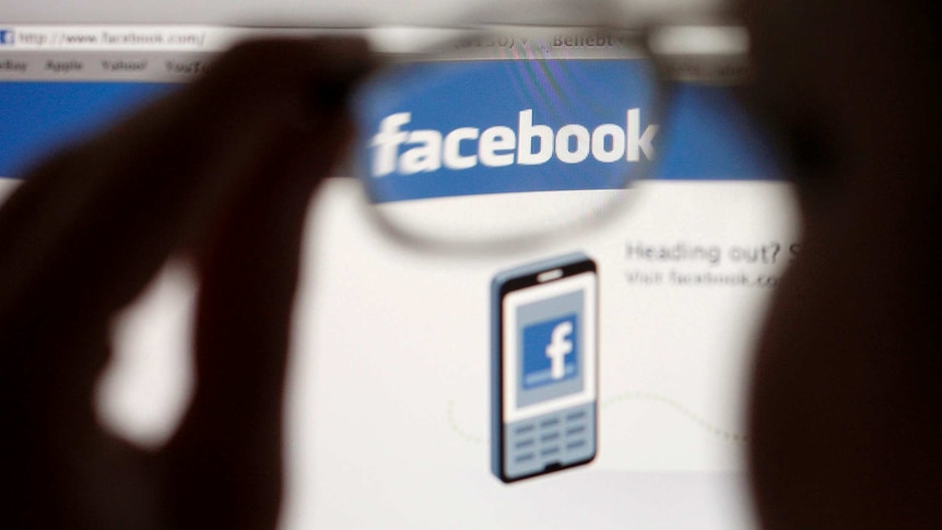 A Facebook logo on a computer screen is seen through glasses held by a woman.