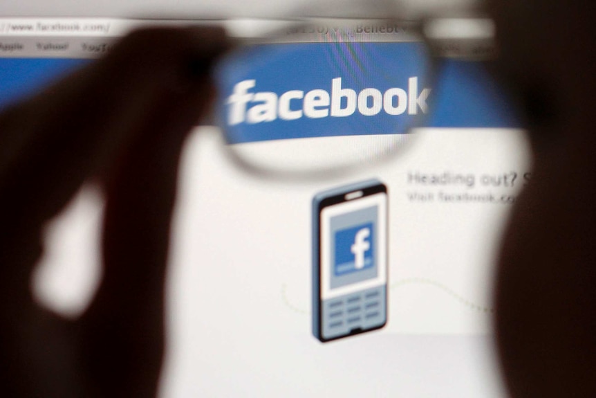 A Facebook logo on a computer screen is seen through glasses held by a woman.