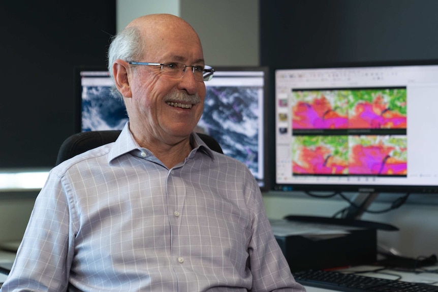 A smiling Mike Bergin sits at the Perth BOM office, with weather maps on computer screens in the background.