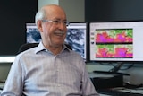 A smiling Mike Bergin sits at the Perth BOM office, with weather maps on computer screens in the background.