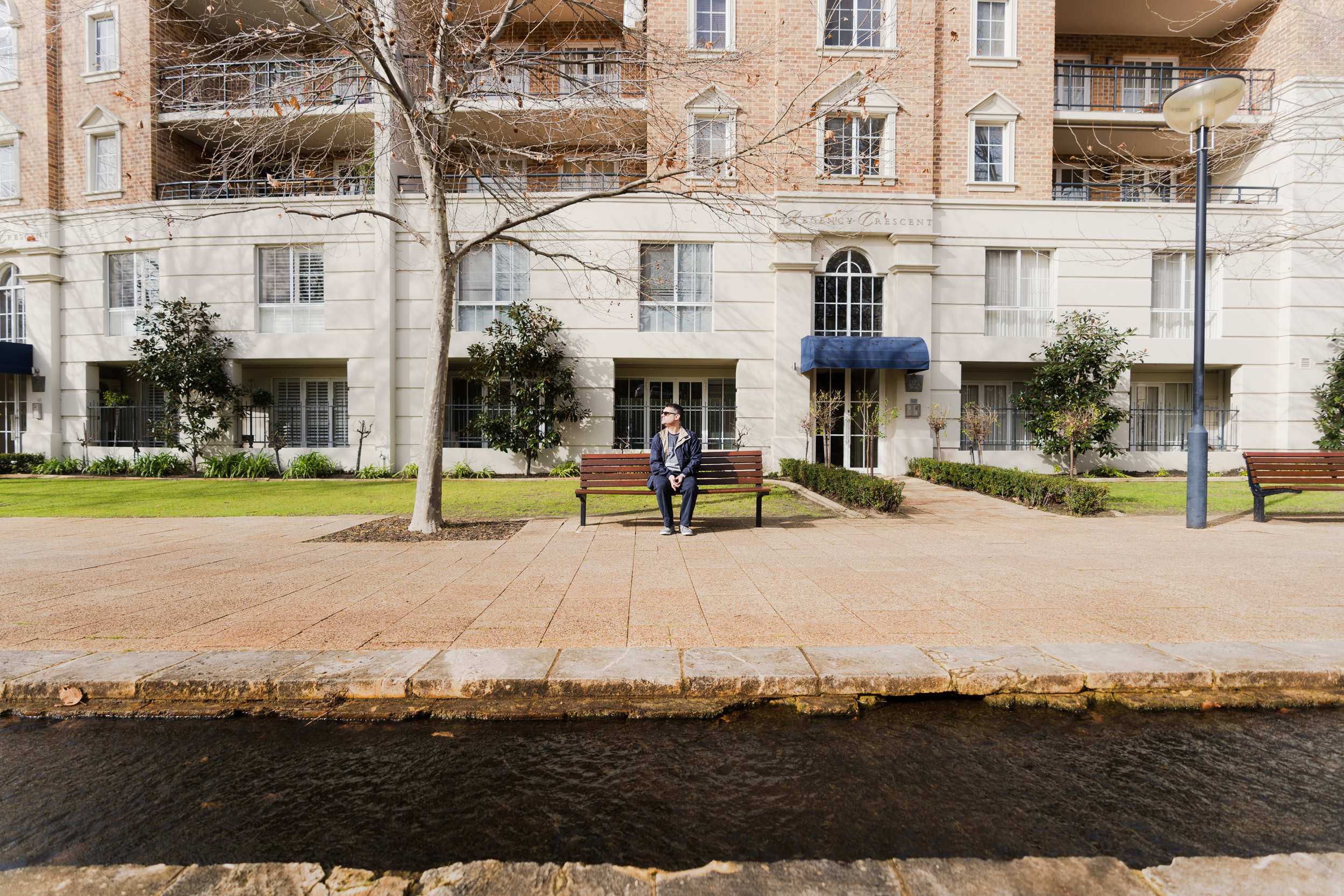 A man with glasses and a navy jacket and pants sits alone on a park bench.