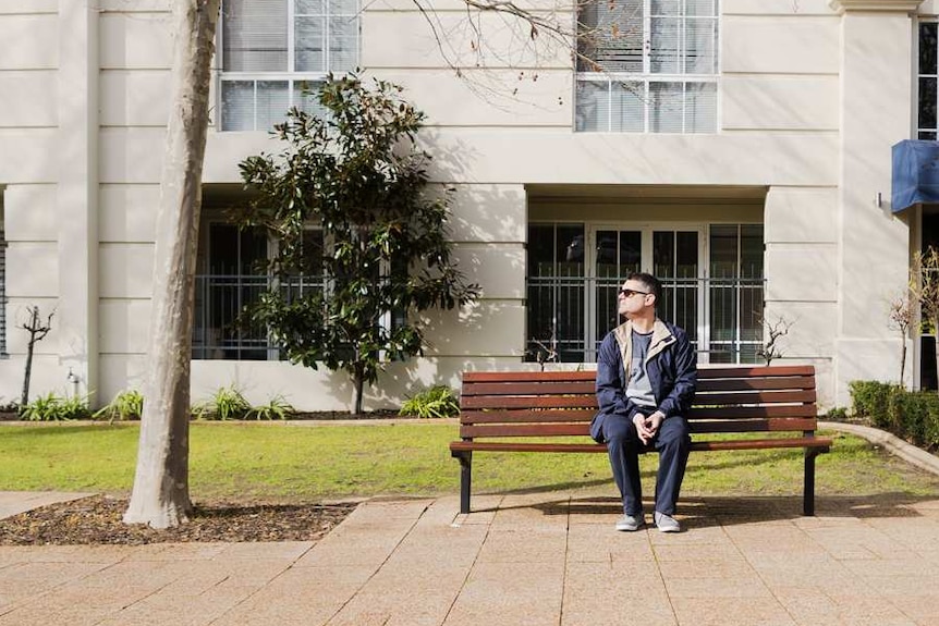 A man with glasses and a navy jacket and pants sits alone on a park bench.
