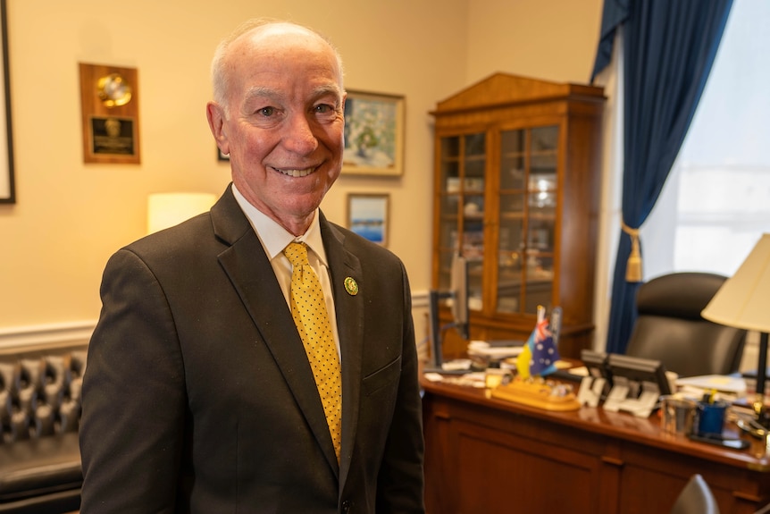 Joe Courtney stands in an office. A desk behind him has a small Australian flag on it.