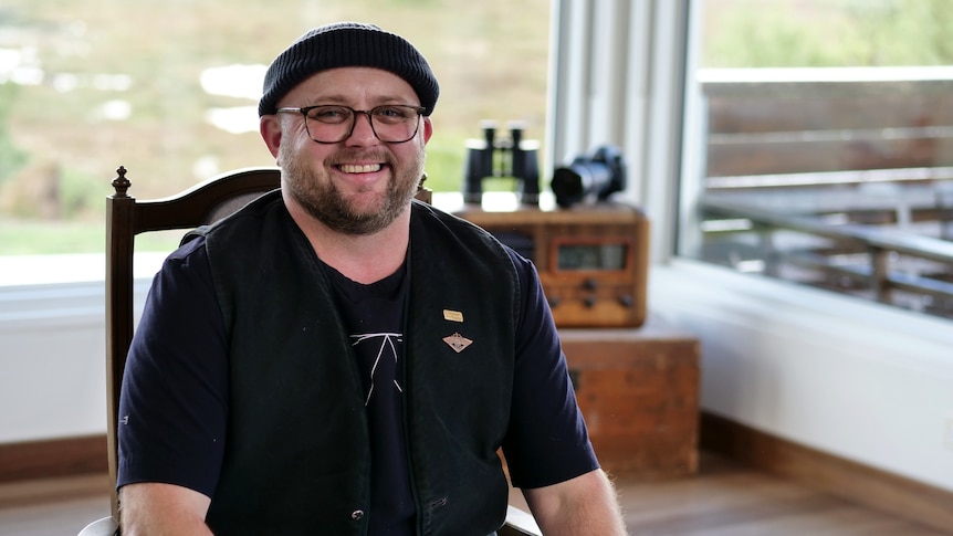 James is sitting in a wooden chair. He is wearing a black beanie and reading glasses and smiling