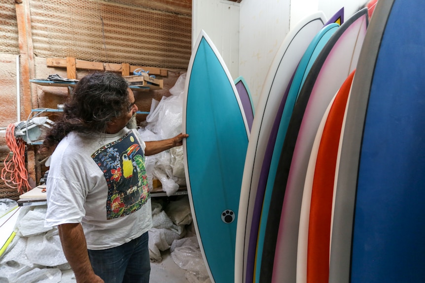 A man examines one of a collection of surfboards mounted in a row inside a large shed.