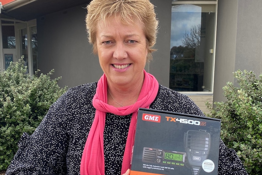 A woman leans on a stone fence, holding an unpoened U-H-F radio in a box.