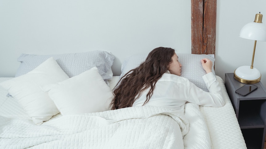 A woman with long brown hair sleeping on her stomach in a large bed made up in white linen