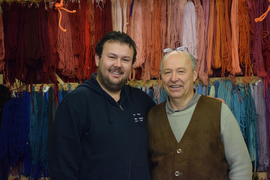 A dark-haired man stands beside his dad in front of a wall upon which threads are hanging. Both are smiling.