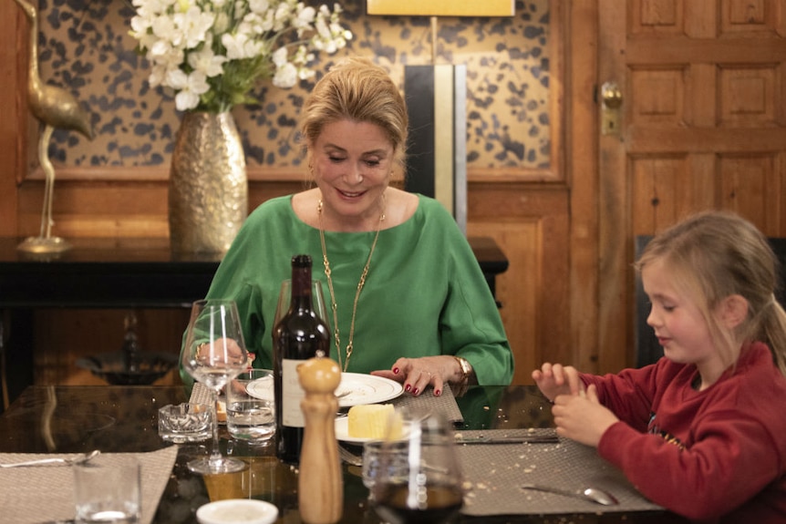 Catherine Deneuve and young girl Clementine Grenier smiling at the dinner table in a room with grand gold decorations