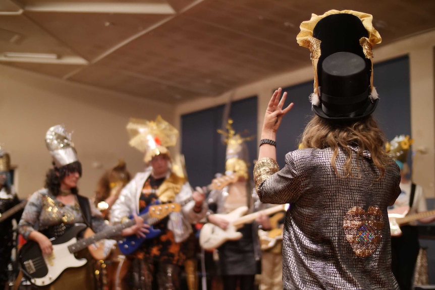 Woman in top hand conducting an all-women band with members wearing shiny costumes, playing in a hall