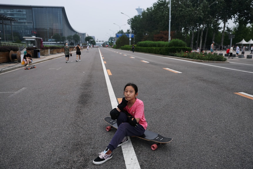 A girl in a pink t-shirt sits in the middle of the road on her skateboard. 