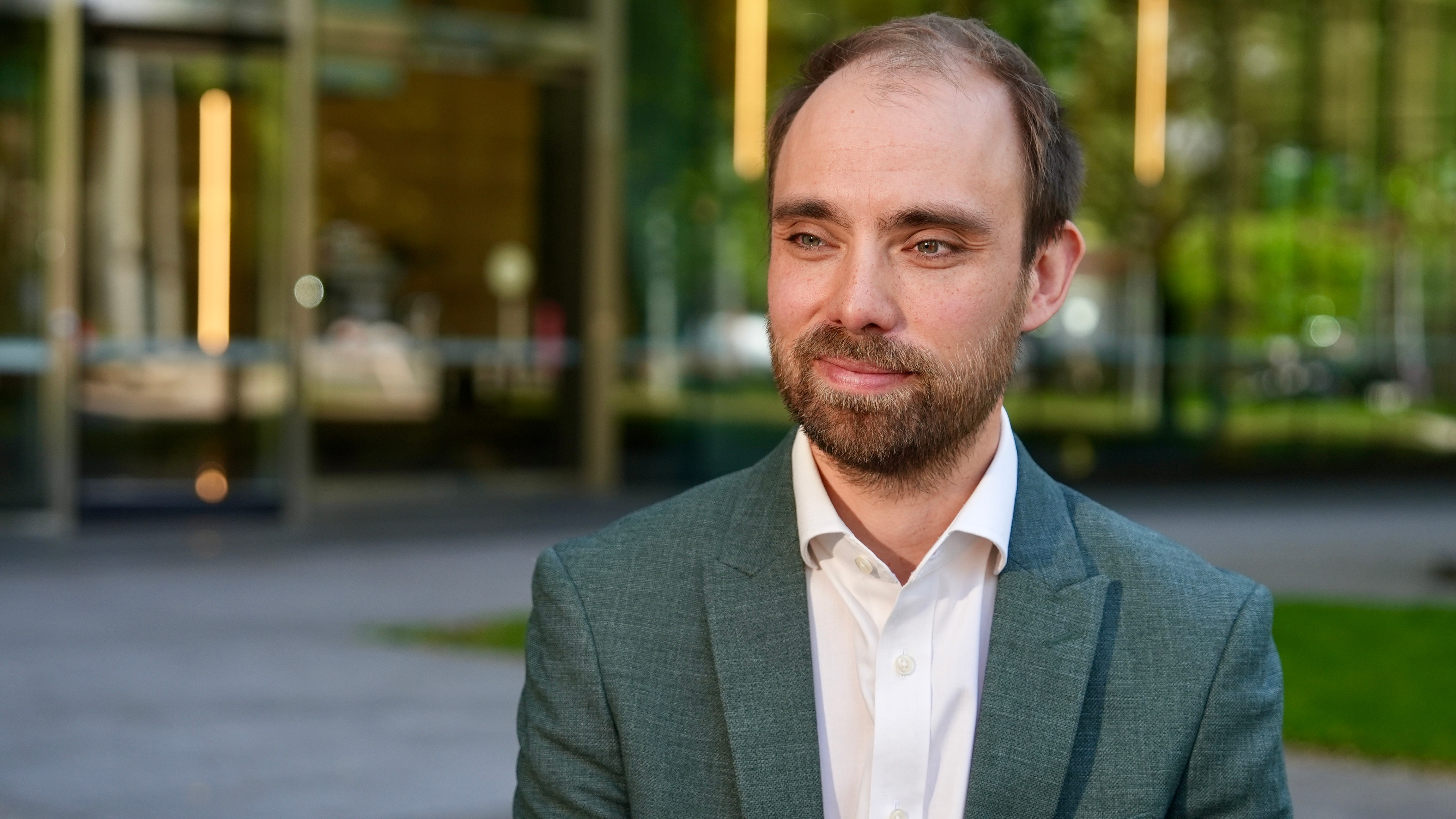 Toby Phillips, wearing a grey suit jacket and a white collared shirt, looks to the side of the camera.