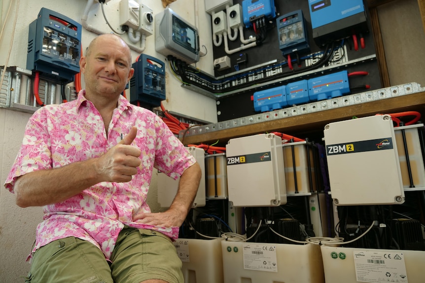 Man in pink floral shirt sitting in a garage, giving a thumbs up with lots of solar and electric equipment behind him
