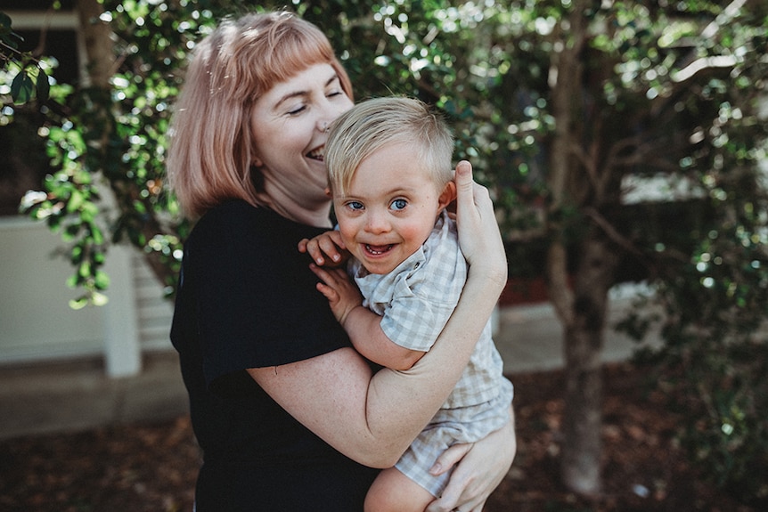 Julia Laine holding young Grayson in her arms in a portrait taken outdoors, both are laughing and smiling.