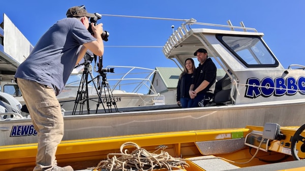 A cameraman films a man and a woman who are both standing in an aluminium fishing boat.
