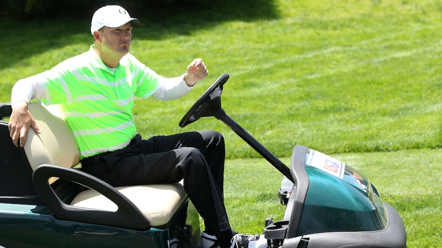 A golfer sits in a golf cart sitting in a fairway at the US Open tournament with his hand just off the steering wheel.