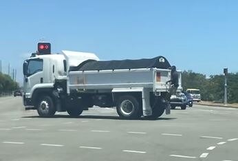 A white tip truck at a traffic light with a man hanging off the back. 