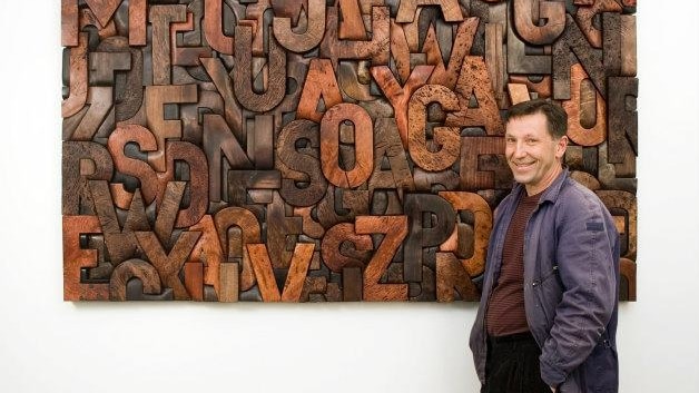 A man stands in front of a large wood carving artwork of jumbled letters