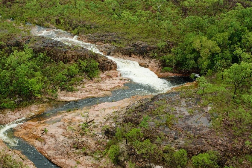An aerial shot of a cascading waterfall, surrounded by green bush land.