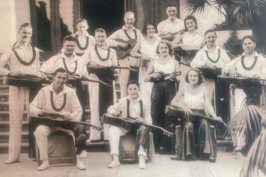 Black and white photo of group of people in matching white outfits and lays, all holding guitars and smiling.