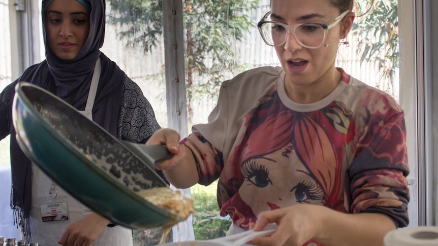 A woman in a headscarf and another in a t-shirt watch mixture pour from a fry pan into a white sieve.