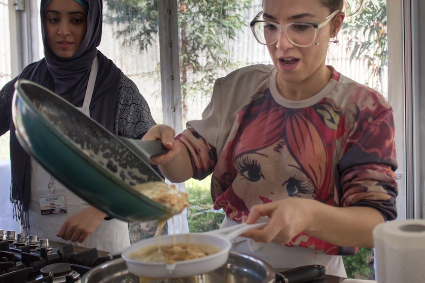 A woman in a headscarf and another in a t-shirt watch mixture pour from a fry pan into a white sieve.