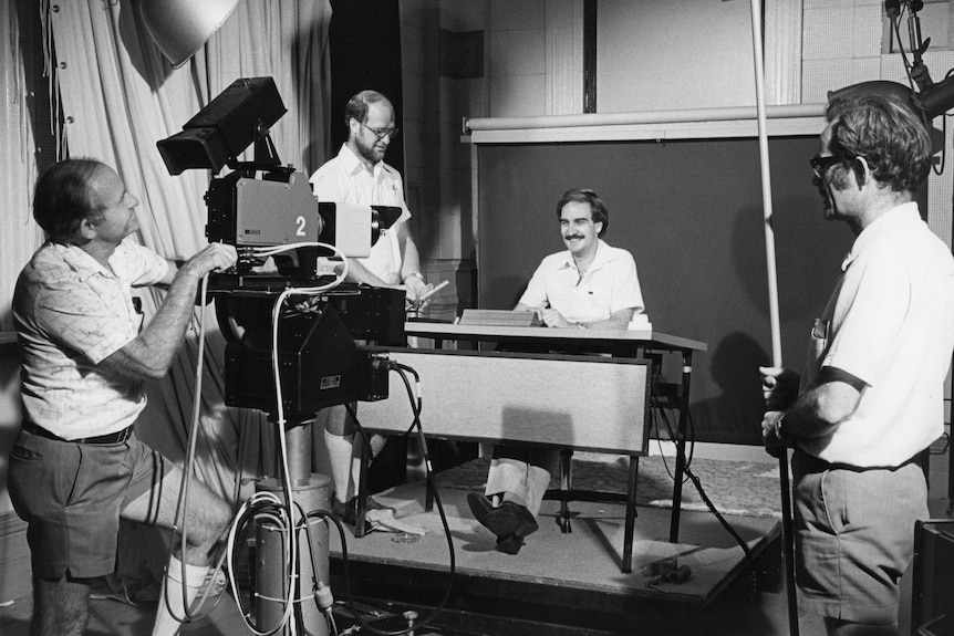 Four men, one behind a big TV camera, one standing to the side and two at a desk filming, black and white.