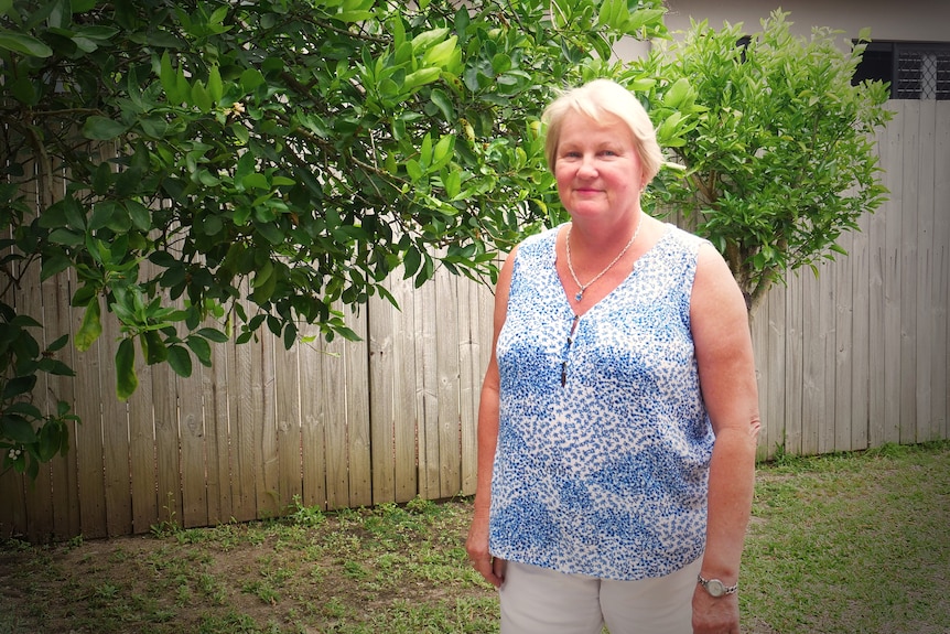 woman judy thatcher stands in yard in front of lime tree and wooden fence wearing blue shirt white pants