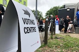 A voting centre sign points to a polling booth where people are lined up waiting to vote.