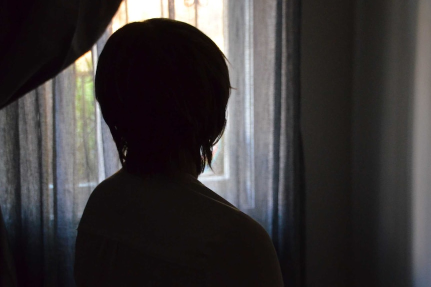 A woman in a wig poses in a shadow in front of a window