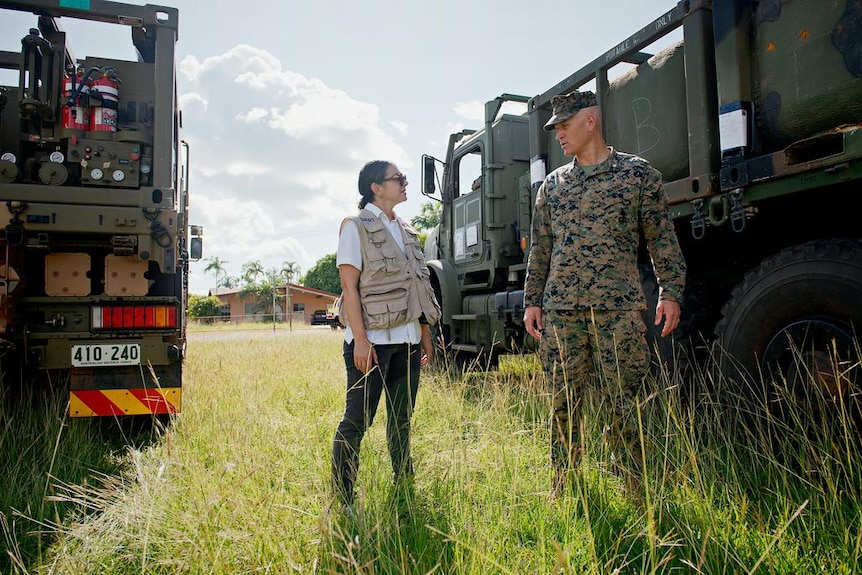 A woman in an army vest talking to a taller US marine as they stand in some grass between two army trucks.