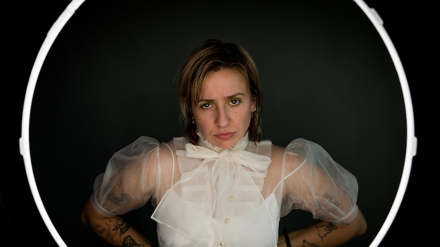 Woman staring at camera with whiet ring around her. Wearing white sheer top. has shoulder length hair.