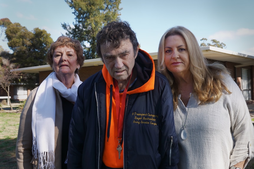 Two women flank a man wearing an army surplus jacket and a bright orange hoody, who is unsmiling. 