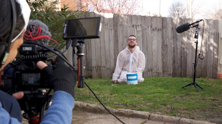 A young man wearing a white suit to protect clothes from paint kneels on the grass by a fence with a camera crew filming him.