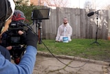A young man wearing a white suit to protect clothes from paint kneels on the grass by a fence with a camera crew filming him.
