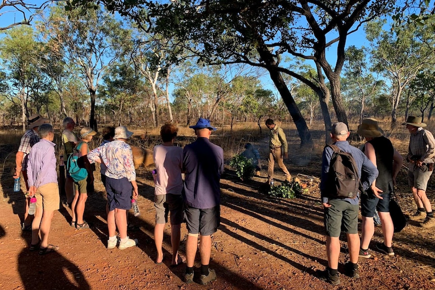 A group of people stand in a circle watching a man light a fire in a smoking ceremony.