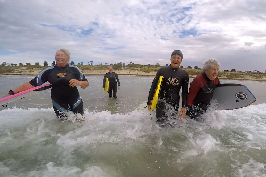 Four retired women wearing wetsuits and holding boogie-boards walk out into the surf to catch more waves