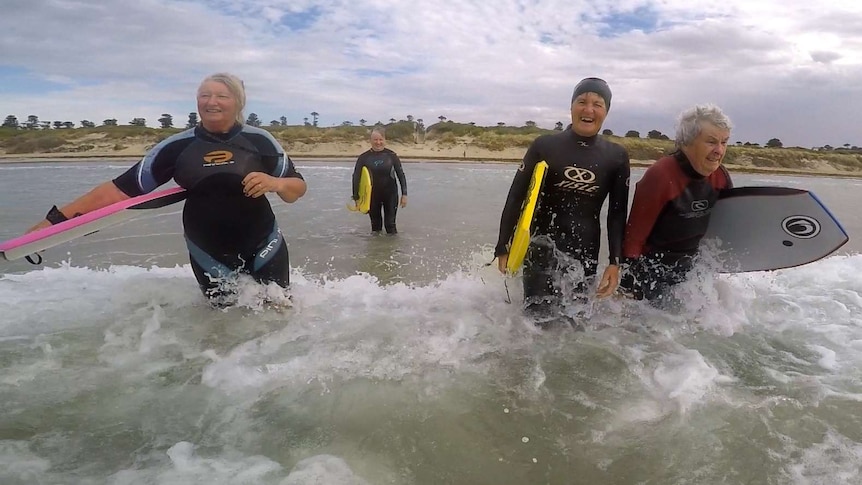 Four retired women wearing wetsuits and holding boogie-boards walk out into the surf to catch more waves