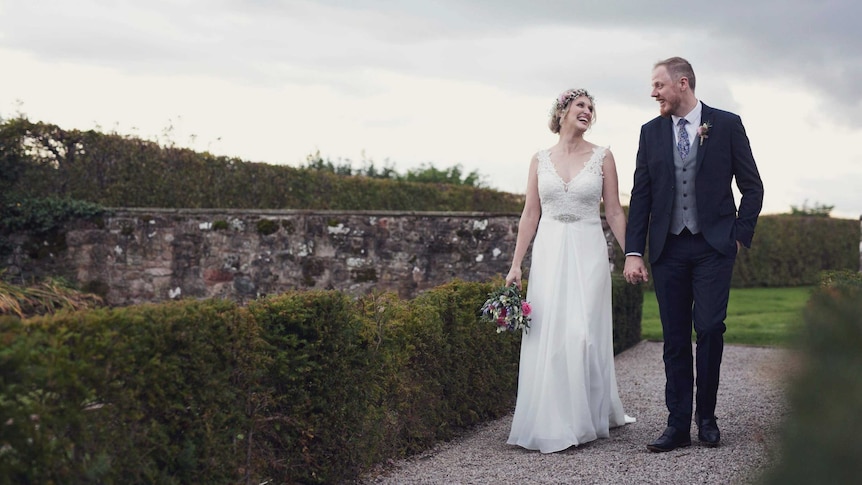A bride and a groom walk hand-in-hand through lush British countryside.