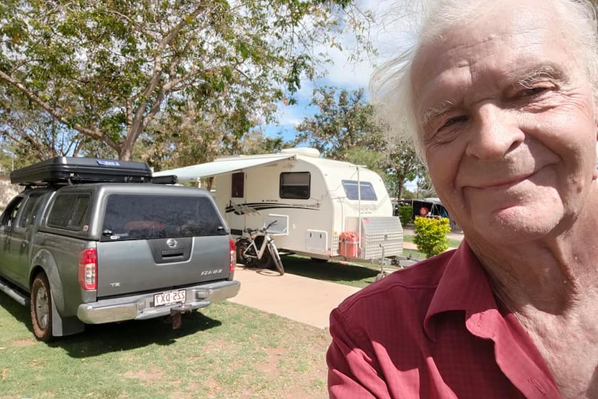 An older gentleman with white hair takes a selfie in front of a grey ute and white caravan.