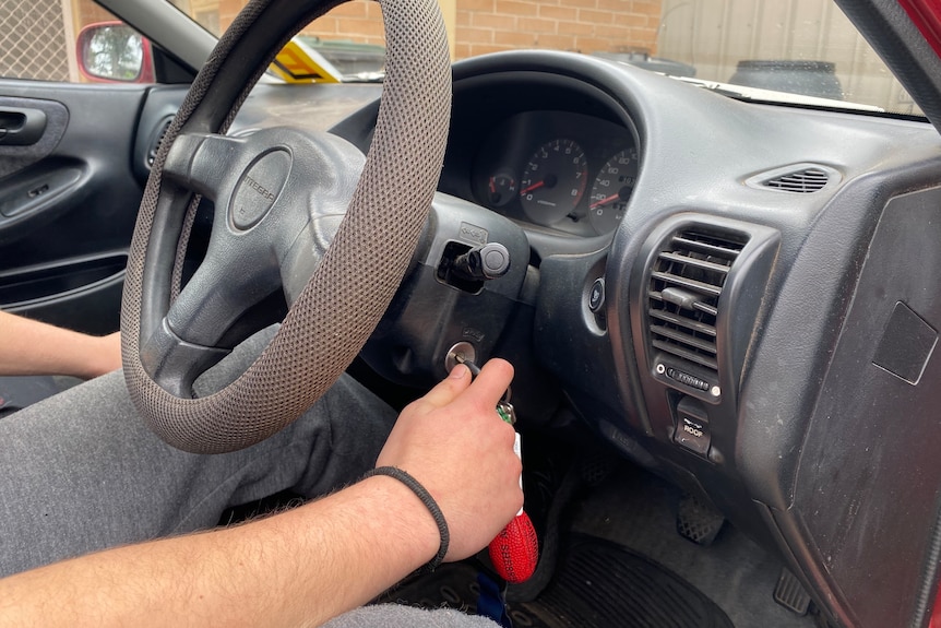 A male hand holds a key in ignition of a vehicle with dark grey interior. He wears grey pants. red p plate is visible on car