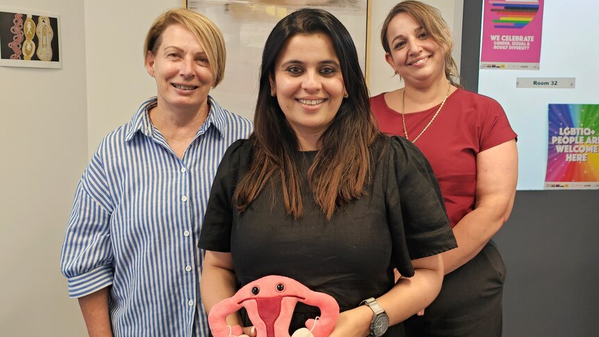 Three women in a medical clinic, the woman in the middle is holding a toy version of the female reproductive system.