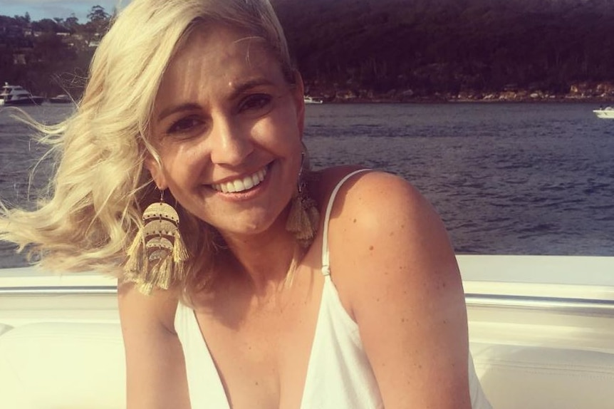 Amity Dry smiles as she sits on a white boat, wearing a white dress and large gold earrings.