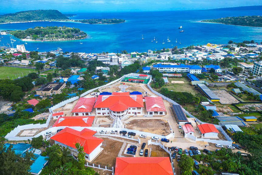 An aerial shot shows a large complex of buildings with red roofs, including a palace, with a bay in the background.