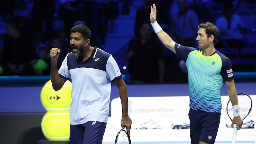 Two men's doubles tennis player react after winning a match, one punching the air and the other waving at the crowd.
