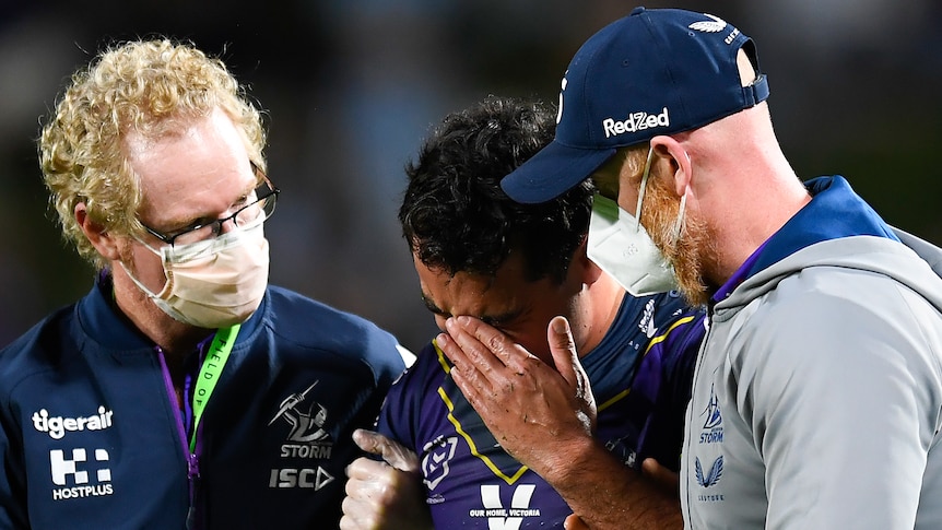 A rugby league player wearing purple holds his hand to his head surrounded by doctors