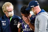 A rugby league player wearing purple holds his hand to his head surrounded by doctors